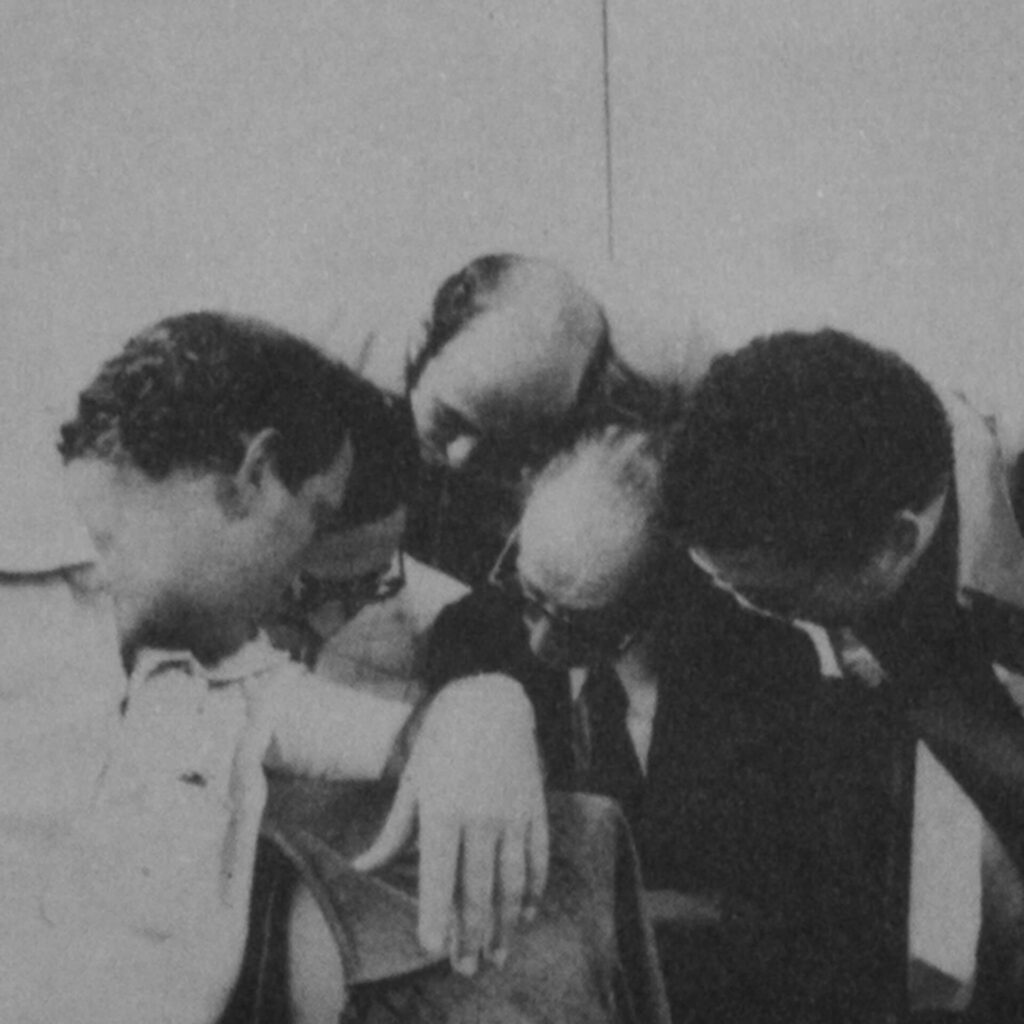 Foto de uma fotografia antiga em preto e branco na vertical. Em uma sala, um
homem branco, calvo, de óculos escuros, terno e gravata está rodeado por outros
quatro homens. Todos olham para baixo e para a esquerda, e parecem conversar
sobre o que veem.